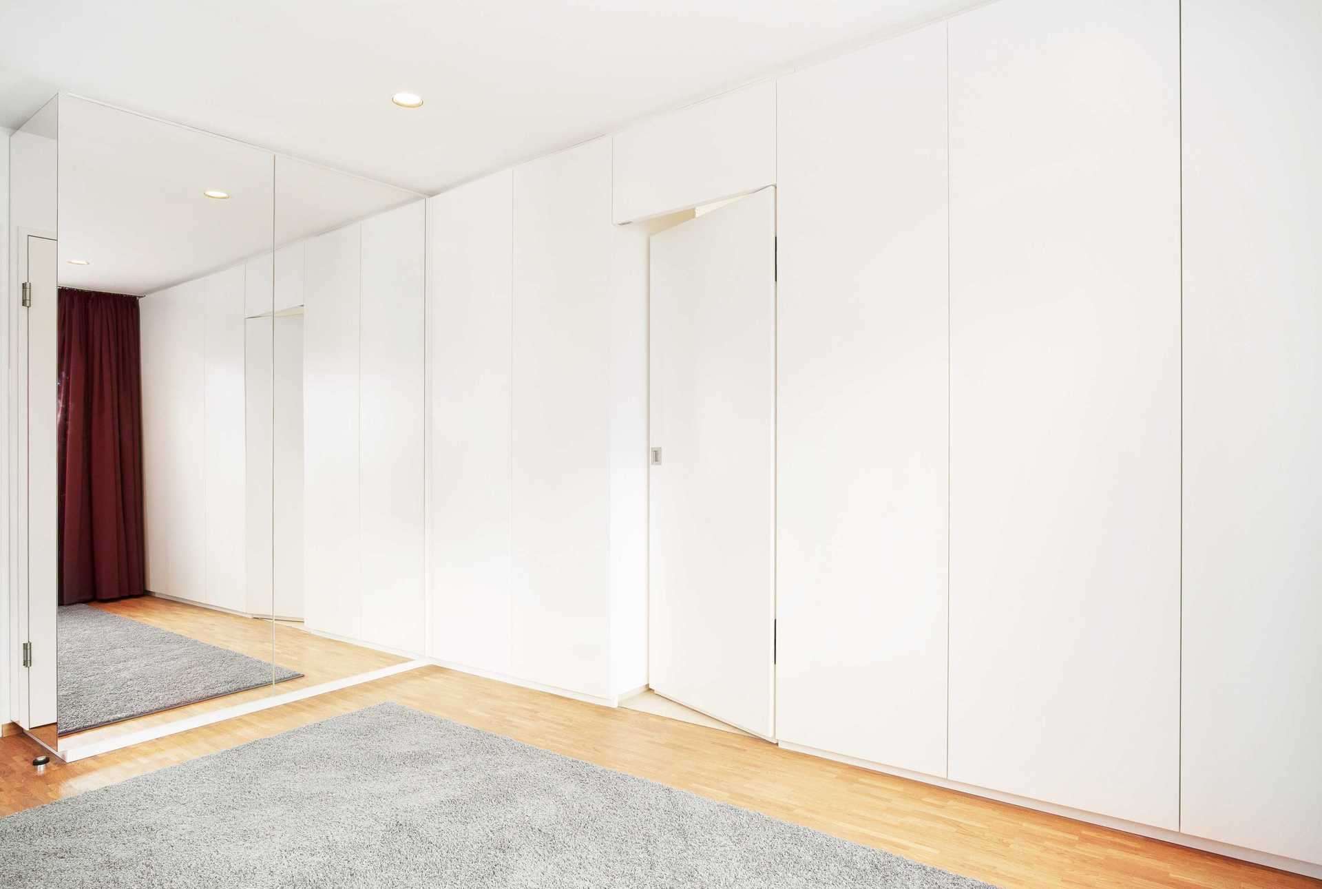 Ankleide/Kleiderschrank über die ganze Wand mit integrierter Tür. Alles in weiß Lack und grifflos. Wandgroßer Spiegel daneben.
