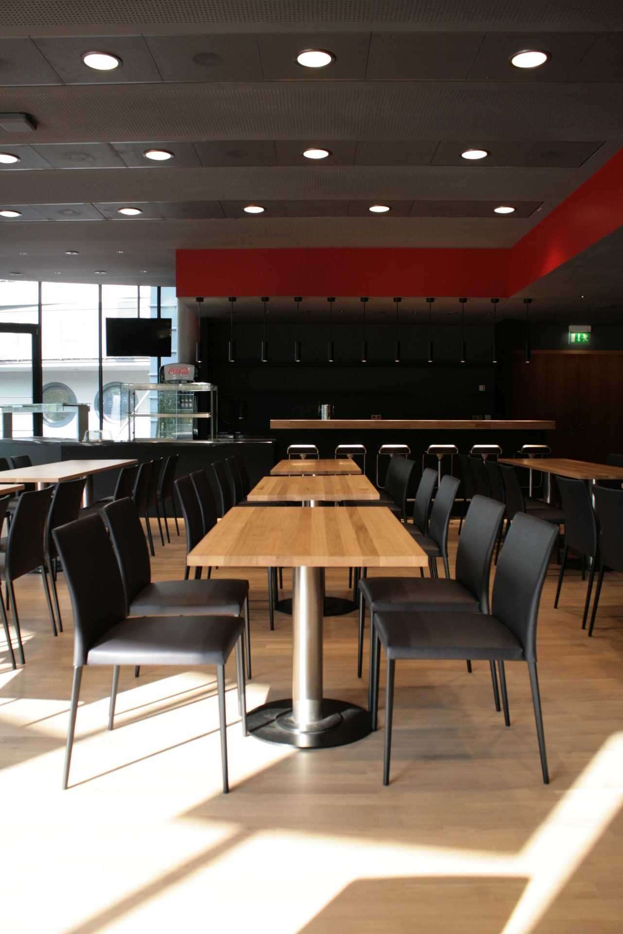 Esstische im Businessbereich des VfB Stuttgarts. Eiche Tische mit schlichten schwarzen Stühlen. Rotes Farbband an der Decke.