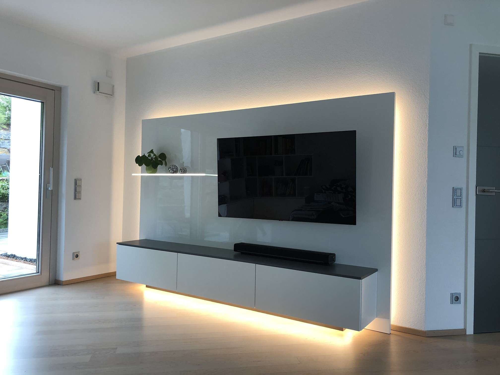 Fernsehmöbel in Glanzlack weiß mit indirektem Licht außenrum, das von hinten strahlt. Rückwand mit niedrigem Sideboard für Stauraum.