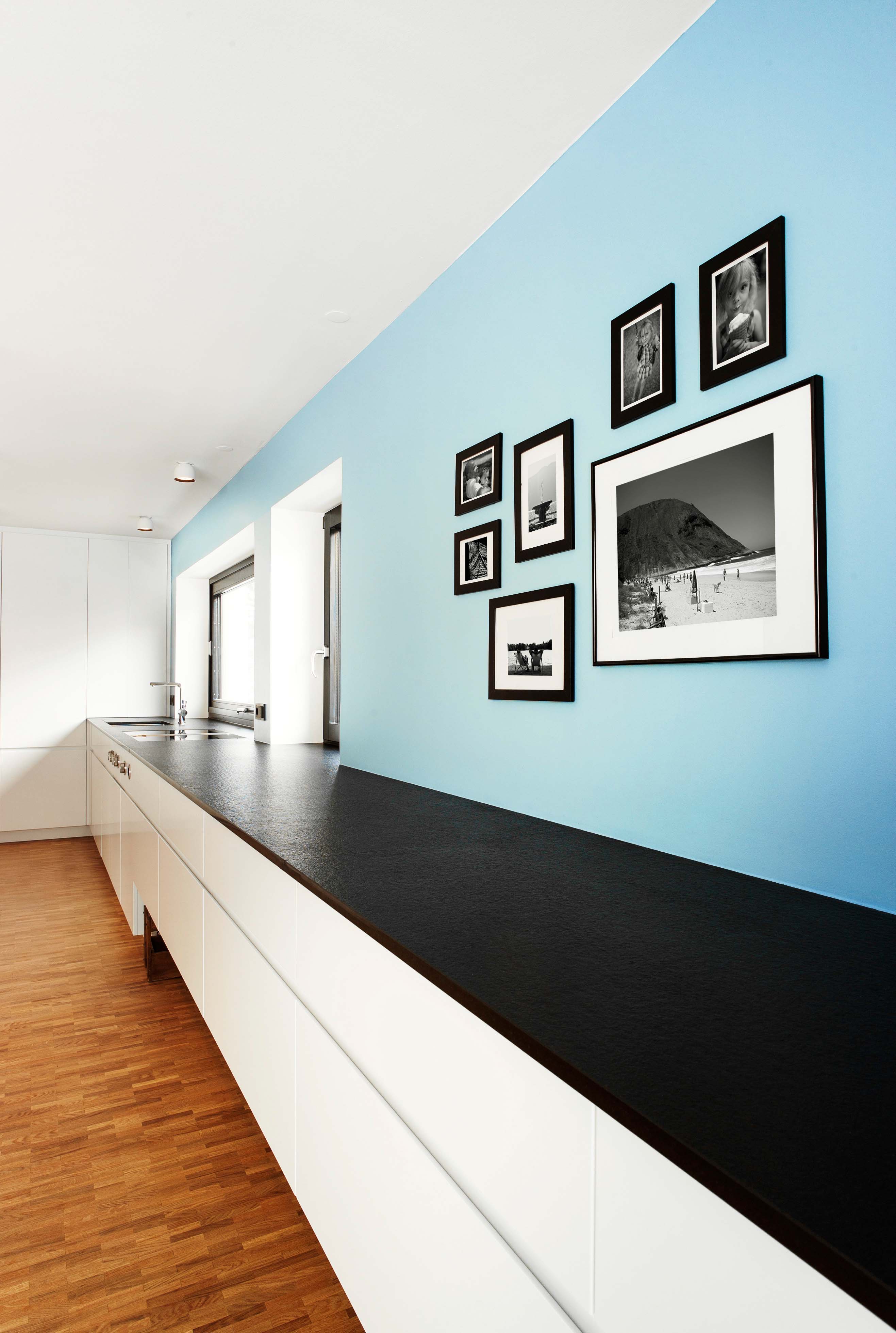 Lange, geradlinige Küchenzeile in weiß, grifflos und mit schwarzer Stein-Arbeitsfläche. Hellblaue Wand dahinter an der Bilder hängen.